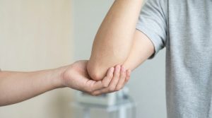 Une main soutient le coude plié d'un patient en T-shirt gris, illustrant un examen médical pour une douleur au bras.